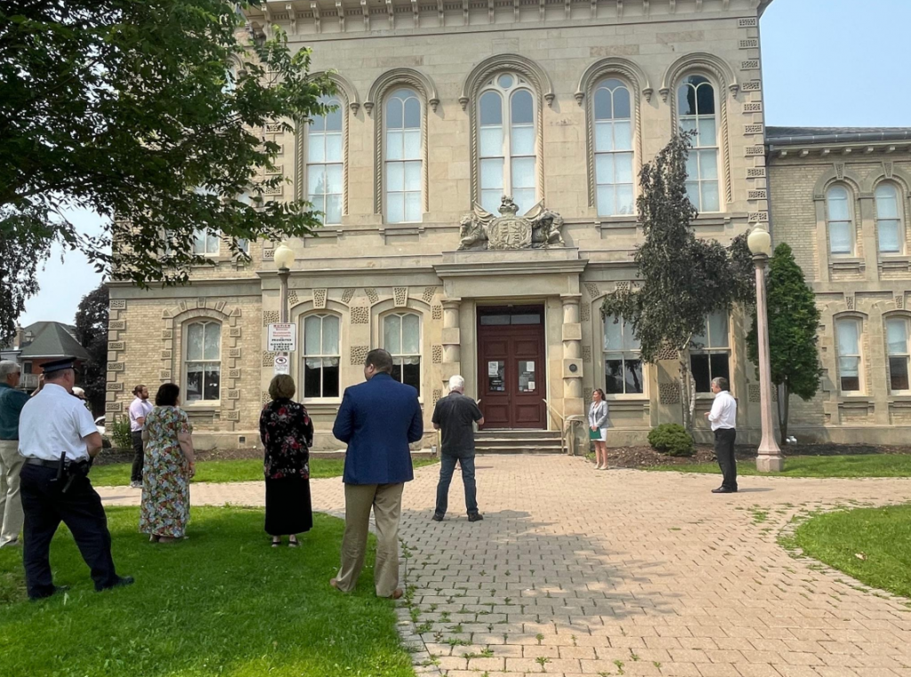 Haldimand-Norfolk Board of Health Chair Kristal Chopp stands in the middle of Governor Simcoe Square, with the Norfolk County Administration Building in the backgroup. An audience of socially distanced staff and community stakeholders are pictured in the foreground, listening to Board Chair Chopp's announcement regarding the finalization of Haldimand and Norfolk's Community Safety & Well-Being Plan. 